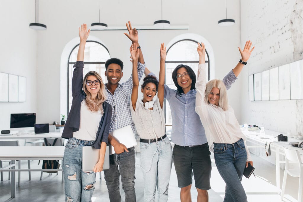 Group of young adults in a casual office setting celebrating with hands in the air
