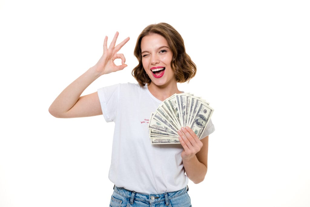 Smiling girl holding bunch of money banknotes and showing ok