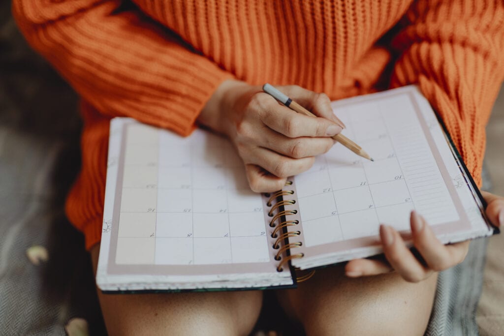 Woman wearing orange sweater writing in planner with a pencil.