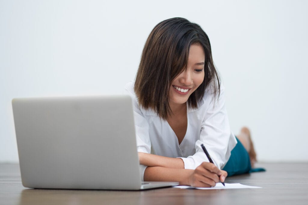 Smiling asian woman writing on floor with her laptop